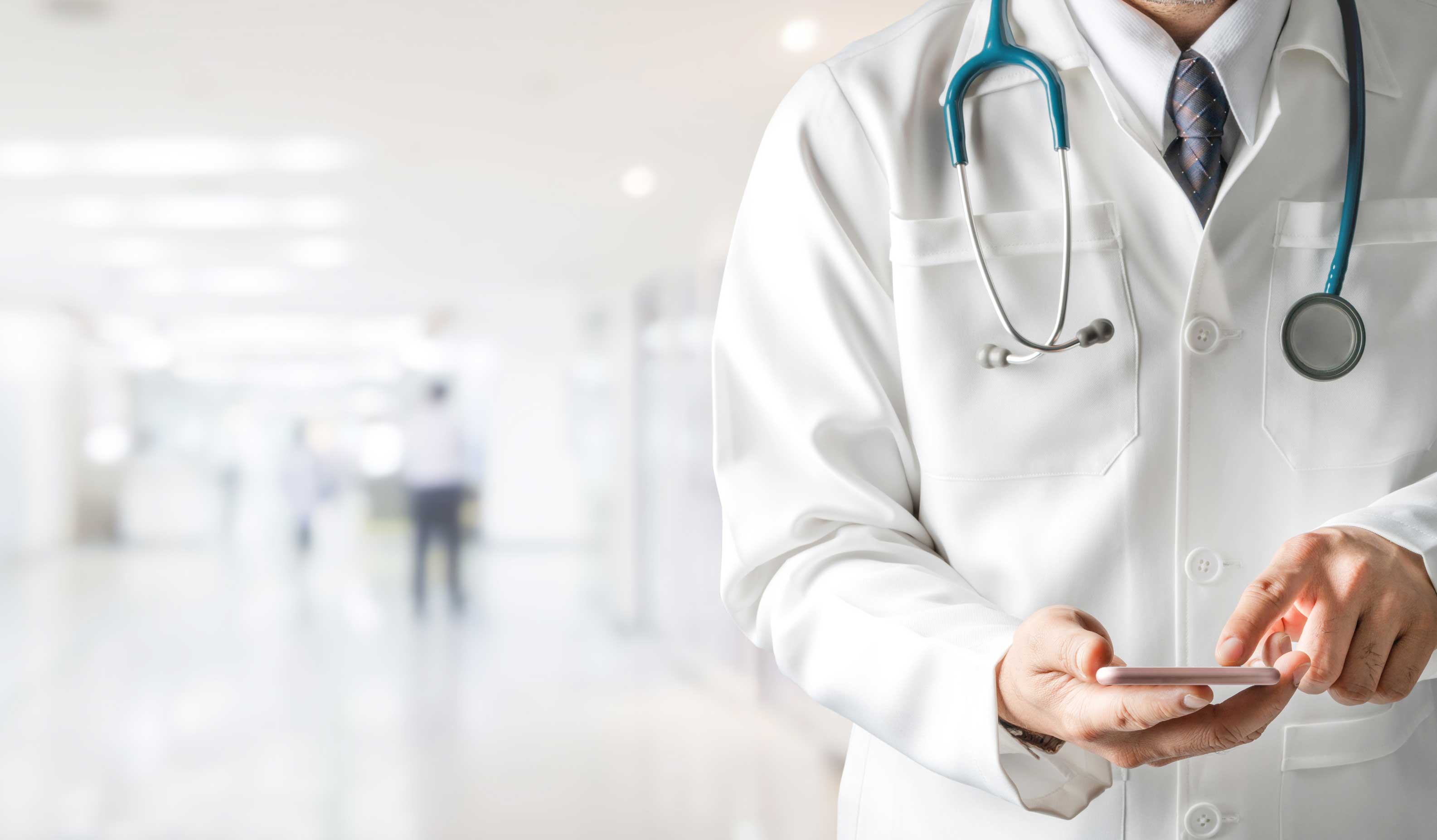 Doctor with stethoscope around neck and cell phone in hand stands in a hospital hallway. Image is cropped so we don't see his face, but can see blurry outlines of other doctors in the background. 