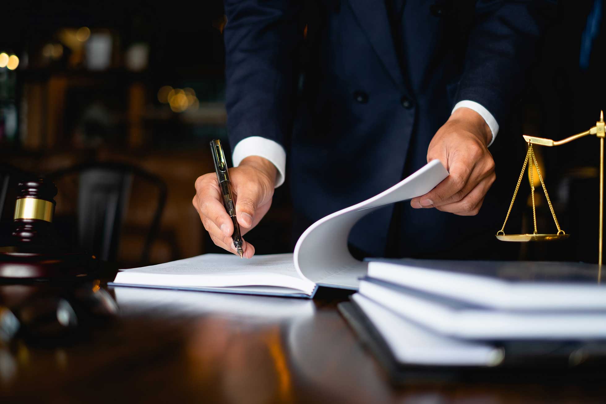 Close up image of a lawyers hands on their desk signing paperwork, a scale and gavel on either side of the image