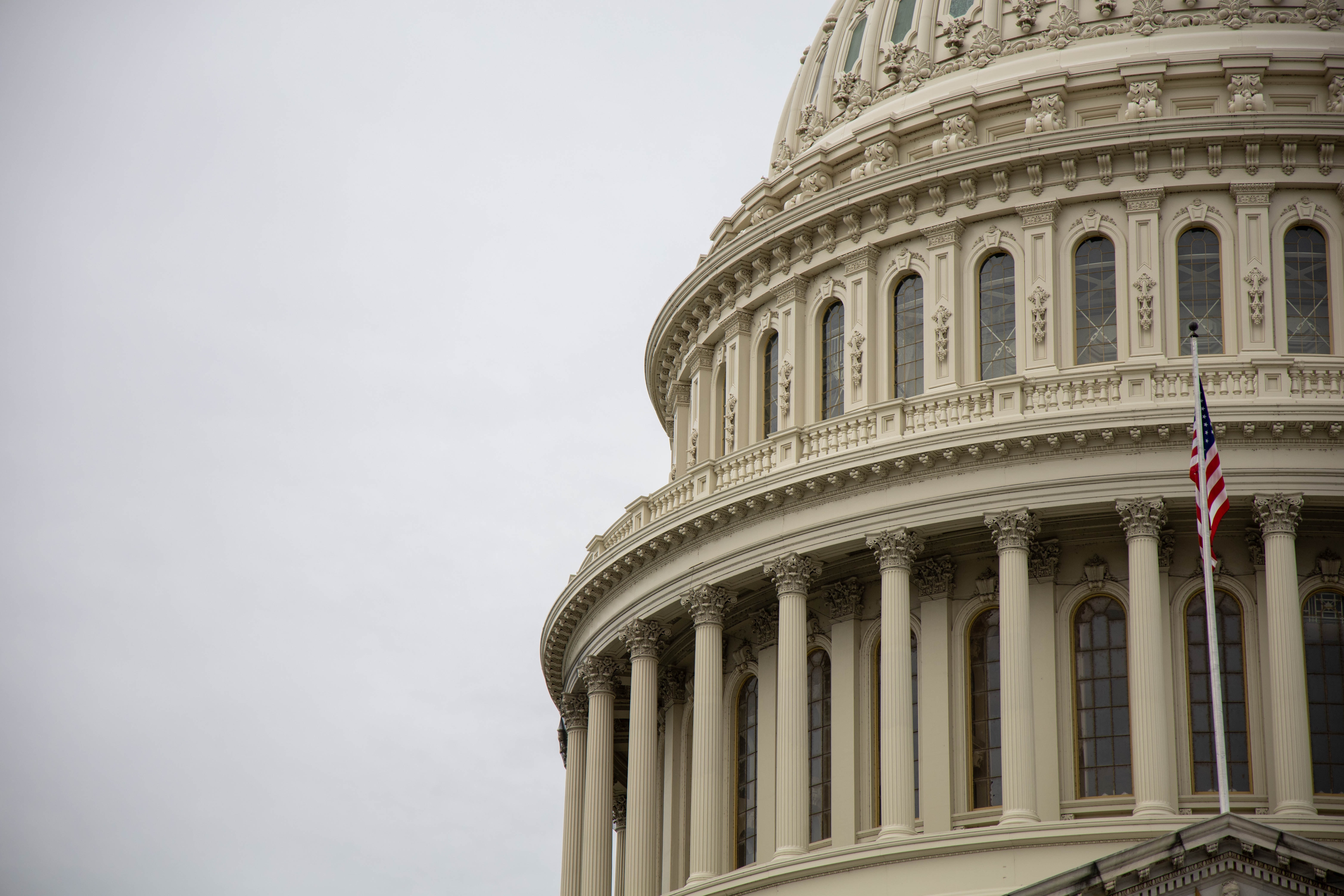 Photo of the top of the dome of the U.S. Capitol building highlighting the architectural details and columns with an American flag on a pole in the foreground. 